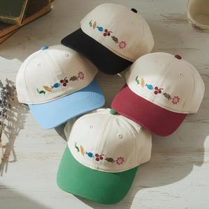 May include: A collection of baseball caps with off-white crowns and colorful brims. Each cap features an embroidered design of leaves, fish, berries, and flowers. The brims are in various colors, including black, blue, red, and green.