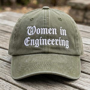 May include: Olive green baseball cap with the text "Women in Engineering" embroidered in white. The cap has a curved brim and a classic six-panel design, suitable for outdoor activities or casual wear.