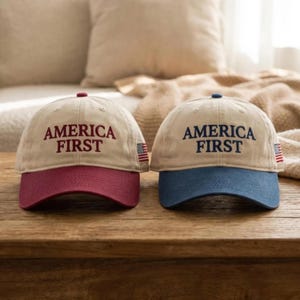 May include: Two baseball caps with the text "AMERICA FIRST" embroidered on the front. One cap has a red brim, the other has a blue brim. Both caps have a beige crown and a small American flag patch.