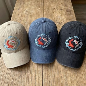 May include: Three baseball caps in tan, blue, and black. Each cap features an embroidered design of two koi fish in red and white, surrounded by blue waves. The caps are made of a soft fabric and have a curved brim.