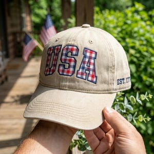 May include: Tan baseball cap with the word "USA" embroidered in red, white, and blue plaid. The cap also features "EST. 177" on the side. The cap is held in front of a blurred background with American flags.