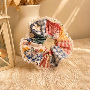May include: A colorful scrunchie with a patchwork design. The fabric features floral, gingham, and striped patterns in shades of blue, red, yellow, and green. The scrunchie is trimmed with white lace and is a hair accessory.