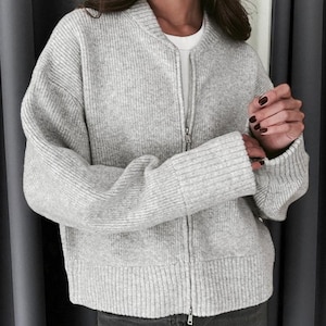 Peut inclure: Veste de bombardier en tricot c&ocirc;tel&eacute; gris clair avec une fermeture &eacute;clair sur toute la longueur. La veste a un col montant et des poignets c&ocirc;tel&eacute;s. Le mod&egrave;le porte un t-shirt blanc et un jean gris fonc&eacute;.