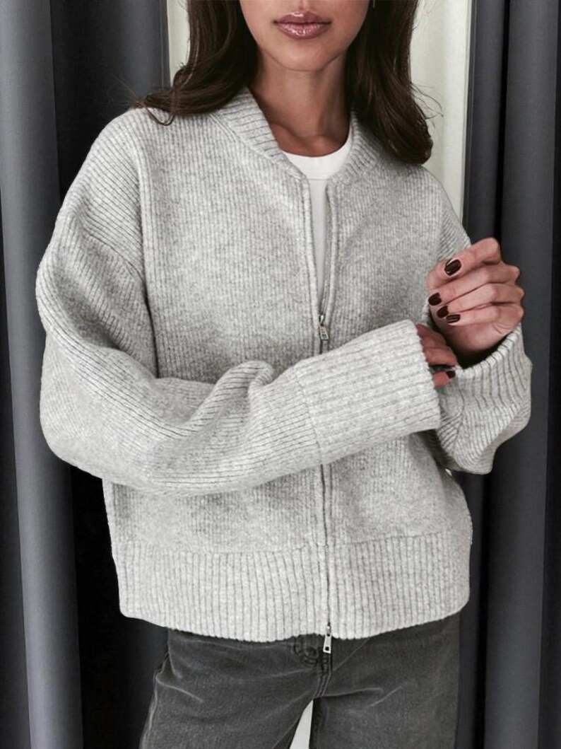 Peut inclure: Veste de bombardier en tricot c&ocirc;tel&eacute; gris clair avec une fermeture &eacute;clair sur toute la longueur. La veste a un col montant et des poignets c&ocirc;tel&eacute;s. Le mod&egrave;le porte un t-shirt blanc et un jean gris fonc&eacute;.