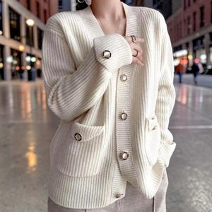 Puede incluir: Cárdigan de punto grueso color crema con cuello en V, bolsillos y botones dorados decorativos. El suéter de manga larga presenta puños acanalados con detalles de botones dorados a juego. Una prenda de abrigo elegante y cálida.