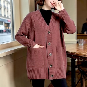 May include: A long, button-down cardigan in a muted rose color. The sweater features a V-neck, two front pockets, and ribbed cuffs and hem. The cardigan has a cable knit detail on the upper left sleeve.
