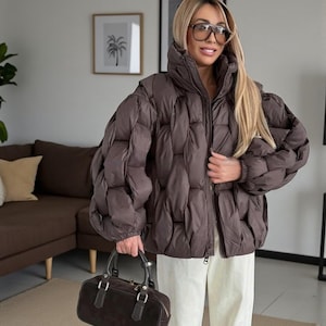 May include: A brown quilted puffer jacket with a high collar and zip fastening. The jacket has long sleeves. A brown handbag with a top handle is also visible. The person is wearing white trousers and glasses.