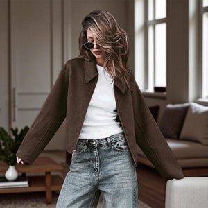 May include: A woman wearing a brown jacket, white shirt, and blue jeans. The jacket has a collared neckline and is open in the front. The jeans are a light wash denim. The background is a living room with a couch and windows.