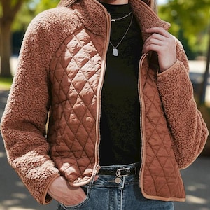 May include: A brown quilted jacket with a high collar and a full zip. The jacket features a quilted front panel and fluffy, textured sleeves. The model is wearing blue jeans and a black top. A warm and stylish outerwear option.