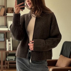 Peut inclure: Un cardigan zipp&eacute; marron fonc&eacute; avec des poignets et un ourlet c&ocirc;tel&eacute;s. Le cardigan est port&eacute; sur un haut de couleur claire et associ&eacute; &agrave; un jean gris. Le cardigan a un col montant et une fermeture &eacute;clair argent&eacute;e.