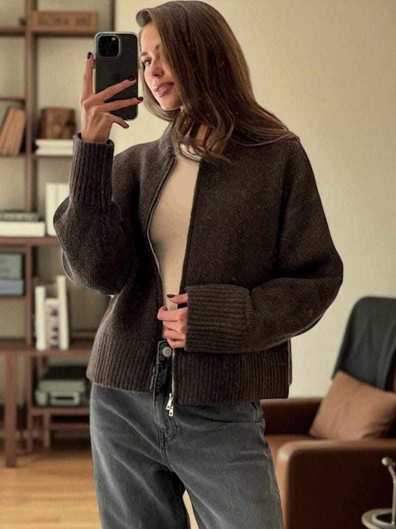 Peut inclure: Un cardigan zipp&eacute; marron fonc&eacute; avec des poignets et un ourlet c&ocirc;tel&eacute;s. Le cardigan est port&eacute; sur un haut de couleur claire et associ&eacute; &agrave; un jean gris. Le cardigan a un col montant et une fermeture &eacute;clair argent&eacute;e.
