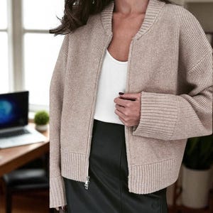Peut inclure: Veste beige en tricot c&ocirc;tel&eacute; avec fermeture &eacute;clair et col montant. Port&eacute;e sur un d&eacute;bardeur blanc et une jupe en cuir noire. La veste a une fermeture &eacute;clair et des poignets c&ocirc;tel&eacute;s.