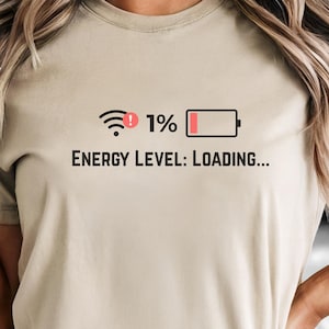Puede incluir: Camiseta color beige con la frase "Energy Level: Loading..." y un gráfico de batería baja y un símbolo de Wi-Fi. La camiseta se combina con unos shorts vaqueros grises.