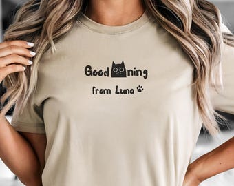 May include: A beige t-shirt with the text "Good morning from Luna" in black. The design includes a cat silhouette and paw prints. The shirt is paired with gray denim shorts.