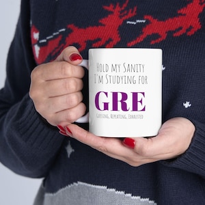 May include: White ceramic mug with the text "HOLD MY SANITY I'M STUDYING FOR GRE" in black and purple. Below the word GRE, the text reads "GUESSING, REPEATING, EXHAUSTED". The mug is held by a person wearing a dark blue sweater.