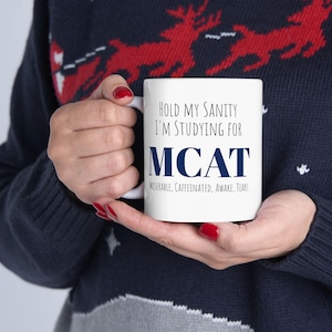 May include: White ceramic mug with the text "HOLD MY SANITY I'M STUDYING FOR MCAT" in black and navy blue. The bottom of the mug reads "MISERABLE, CAFFEINATED, AWAKE, TEARS". The mug is held by a person wearing a navy blue sweater.