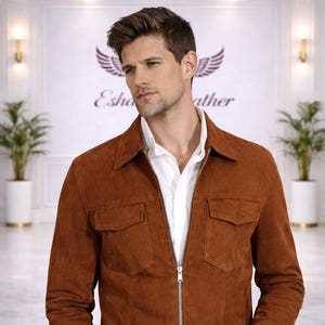 Könnte beinhalten: Braune Wildlederjacke mit Reißverschluss und zwei Vordertaschen. Die Jacke wird über einem weißen Hemd mit Kragen getragen. Der Hintergrund zeigt ein Logo mit dem Text "Esha Leather".
