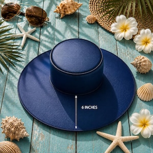 Puede incluir: Un sombrero de sol azul marino con un ala de 15 cm, rodeado de conchas marinas, estrellas de mar y gafas de sol. El sombrero tiene una copa redonda y un ala ancha. Accesorios sobre una superficie de madera azul claro envejecida.