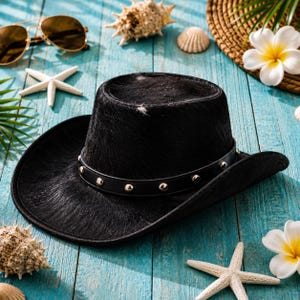 Könnte beinhalten: Ein schwarzer Cowboyhut mit einem Lederband mit Nieten. Der Hut hat eine strukturierte Oberfläche und eine breite Krempe. Der Hut ist umgeben von Muscheln, Sonnenbrillen und tropischen Blumen auf einer blauen Holzoberfläche.