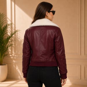 K&ouml;nnte beinhalten: Eine bordeauxrote Leder-Bomberjacke mit wei&szlig;em Kunstfellkragen, langen &Auml;rmeln mit gerippten B&uuml;ndchen und einem gerippten Saum. Die Jacke wird mit einer schwarzen Hose und einer schwarzen Sonnenbrille getragen und zeigt strukturierte N&auml;hte am R&uuml;cken.