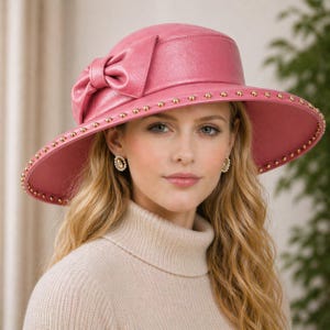 Könnte beinhalten: Ein rosa Lederhut mit breiter Krempe und großer Schleife an der Seite. Der Rand ist mit kleinen, goldfarbenen Nieten verziert. Der Hut ist ein stilvolles Accessoire.