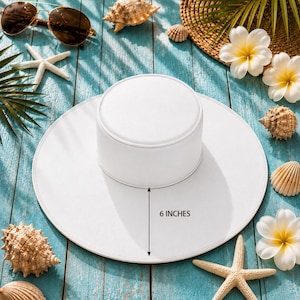 Puede incluir: Un sombrero blanco con una corona de 15 cm, sobre un fondo de playa. La imagen incluye conchas marinas, estrellas de mar, gafas de sol y flores tropicales sobre una superficie de madera azul.