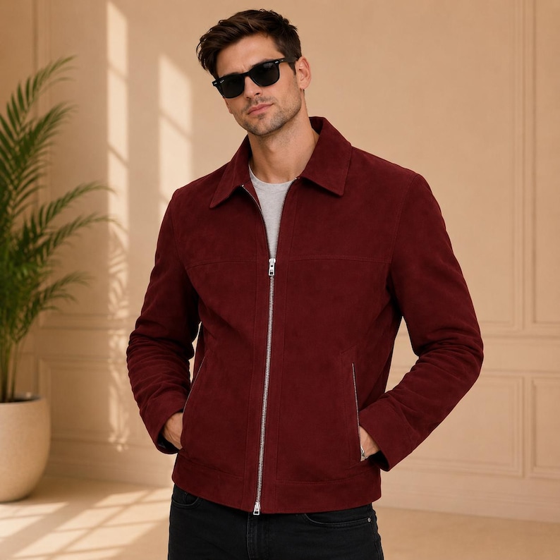 May include: A deep burgundy suede jacket featuring a classic collar, a full-length silver-toned metal zipper closure, and two vertical zippered side pockets. The jacket is worn over a plain light gray t-shirt and paired with dark trousers.
