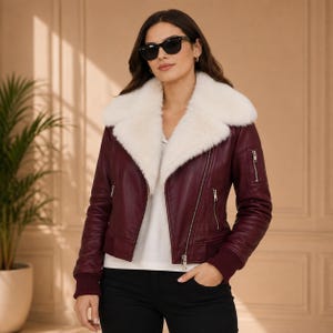K&ouml;nnte beinhalten: Eine tiefburgunderrote Lederjacke mit einem gro&szlig;en, flauschigen wei&szlig;en Kunstfellkragen, silberfarbenen Metallrei&szlig;verschl&uuml;ssen an der Vorderseite und den &Auml;rmeln sowie gerippten Strickb&uuml;ndchen. Die Jacke wird &uuml;ber einem wei&szlig;en Oberteil und einer schwarzen Hose getragen.