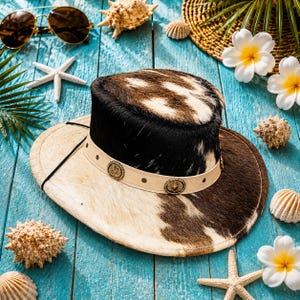 Könnte beinhalten: Ein stilvoller Cowboyhut mit einem einzigartigen Kuhfellmuster in Braun-, Schwarz- und Weißtönen. Der Hut hat ein cremefarbenes Band mit dekorativen Metallakzenten. Muscheln, Sonnenbrillen und tropische Blumen liegen auf einer blauen Holzoberfläche.