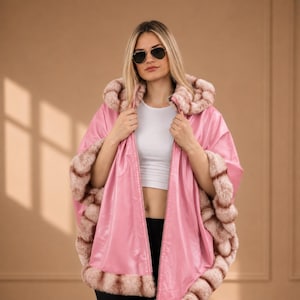 Könnte beinhalten: Ein rosa Kapuzenponcho mit Kunstfellbesatz und -futter. Der Poncho hat einen Reißverschluss und wird über einem weißen Oberteil getragen. Das Model trägt eine Sonnenbrille.