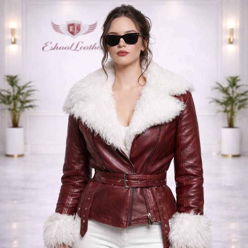 K&ouml;nnte beinhalten: Burgunderfarbene Lederjacke mit wei&szlig;em Pelzkragen und Manschetten. Die Jacke hat einen G&uuml;rtel in der Taille und einen Rei&szlig;verschluss. Das Model tr&auml;gt eine Sonnenbrille und eine wei&szlig;e Hose. Der Markenname "Eshaal Leather" ist im Hintergrund zu sehen.