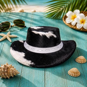 Könnte beinhalten: Ein schwarz-weißer Kuhfell-Cowboyhut mit weißem Band. Der Hut liegt auf einer türkisfarbenen Holzoberfläche mit Muscheln, Sonnenbrille und tropischen Blumen. Der Hut hat eine breite Krempe und eine hohe Krone.
