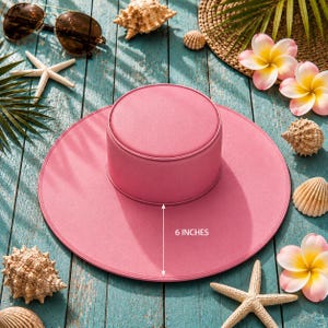 Puede incluir: Un sombrero de sol rosa con una corona de 15 cm, rodeado de conchas marinas, estrellas de mar y gafas de sol sobre una superficie de madera azul. El sombrero tiene un ala ancha y una corona cil&iacute;ndrica.