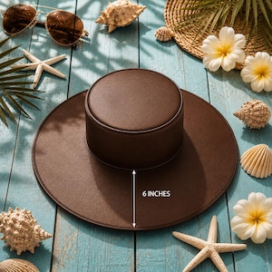 Puede incluir: Un sombrero marr&oacute;n con copa redonda y ala ancha, de 15 cm de altura. El sombrero se exhibe sobre una superficie de madera azul, rodeado de conchas marinas, estrellas de mar y gafas de sol, evocando un tema playero.