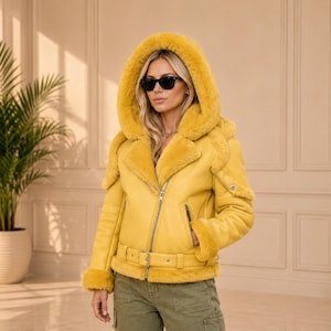 Könnte beinhalten: Eine leuchtend gelbe Lederjacke mit einer großen Kapuze, die mit passendem gelbem Kunstfell gefüttert ist. Die Jacke verfügt über einen silberfarbenen Frontreißverschluss, einen Gürtel mit Schnalle in der Taille sowie Kunstfellbesatz an Ärmeln.