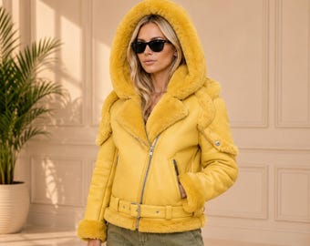 Könnte beinhalten: Eine leuchtend gelbe Lederjacke mit einer großen Kapuze, die mit passendem gelbem Kunstfell gefüttert ist. Die Jacke verfügt über einen silberfarbenen Frontreißverschluss, einen Gürtel mit Schnalle in der Taille sowie Kunstfellbesatz an Ärmeln.