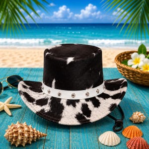 May include: Cowboy hat with a black and white cowhide pattern, a white band with silver studs, and a black chin strap. The hat is on a blue wooden surface with seashells, sunglasses, and a basket of flowers in the background.