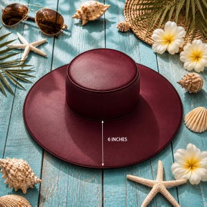 Puede incluir: Un sombrero de sol burdeos con un ala de 15 cm, rodeado de conchas marinas, estrellas de mar y gafas de sol sobre una superficie de madera azul. El sombrero tiene una copa redonda y un ala ancha.