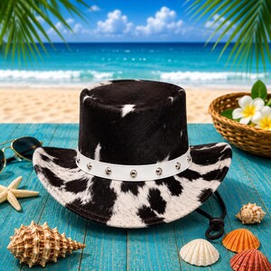 May include: Cowboy hat with a black and white cowhide pattern, a white band with silver studs, and a black chin strap. The hat is on a blue wooden surface with seashells, sunglasses, and a basket of flowers in the background.