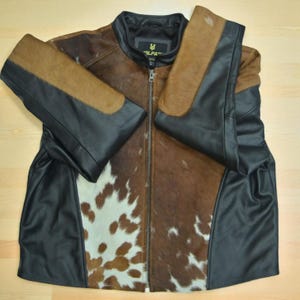 May include: A leather jacket with a cowhide pattern. The jacket features a brown and white cowhide front panel, black leather sleeves and side panels, and a black collar. The jacket has a zipper closure and a small logo on the collar.
