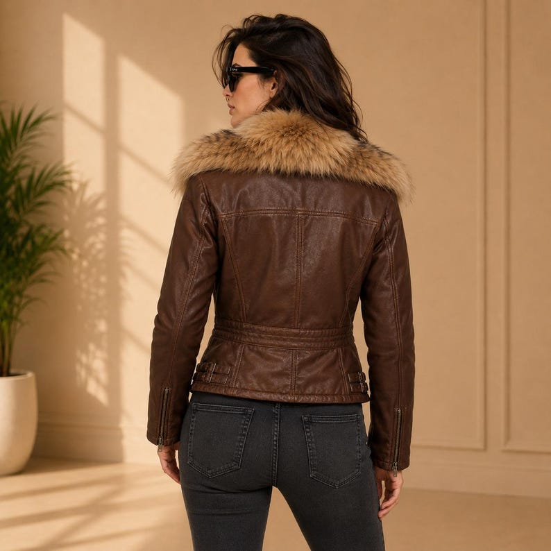 May include: A brown jacket with a tan faux fur collar, long sleeves with zippered cuffs, and side buckle details at the waist. The jacket features visible seam stitching across the back and is paired with dark grey denim jeans.