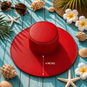 Puede incluir: Un sombrero rojo brillante con una corona de 15 cm, rodeado de conchas marinas, estrellas de mar y gafas de sol sobre una superficie de madera azul. El sombrero es un accesorio elegante para los d&iacute;as soleados.