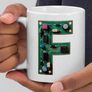 Può includere: Tazza in ceramica bianca con un design a circuito stampato verde a forma di lettera "F". Il circuito stampato include vari componenti elettronici nei toni del verde, rosa e nero. La tazza è tenuta da una persona.