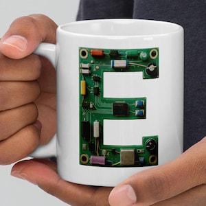 May include: White ceramic mug with the letter "E" formed from a green circuit board design. The mug is held in a person's hands. The design incorporates electronic components in green, red, and yellow, creating a tech-inspired aesthetic.
