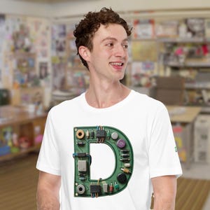 May include: White t-shirt with a large green letter "D" made from electronic components. The design incorporates chips, resistors, and other circuit board elements, creating a unique tech-inspired graphic.