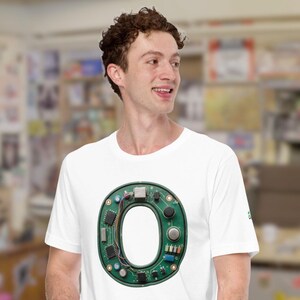 Pode incluir: Camiseta branca com uma grande letra "O" construída a partir de componentes verdes de placa de circuito. O design inclui várias peças eletrônicas, fios e um espaço central aberto formando a letra. A camisa tem um pequeno logotipo verde na manga.