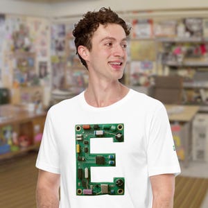 Op de afbeelding: Wit T-shirt met een grote groene printplaat in de vorm van de letter "E". De printplaat heeft diverse elektronische componenten in verschillende kleuren. De persoon draagt een wit T-shirt.