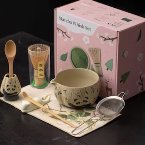 Peut inclure: Un ensemble de préparation de matcha avec une boîte rose illustrant le thé matcha et le bambou. L'ensemble comprend un fouet en bambou, un bol en céramique avec des motifs de panda, une cuillère, une passoire et une cuillère en bambou.