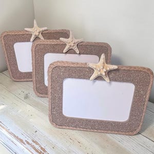 May include: Three rectangular picture frames with rounded corners, featuring a textured tan sand-like surface bordered by braided rope trim. Each frame is adorned with a single, light-colored, five-pointed starfish attached to the top edge.