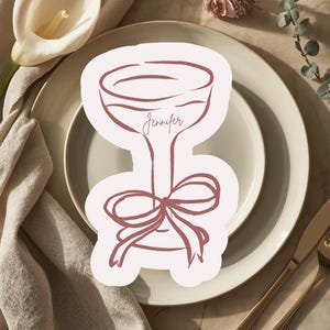 May include: A light pink place card in the shape of a champagne glass, with the name "Jennifer" written in cursive. The place card features a bow at the base and is set on a white plate with gold cutlery and floral decorations.